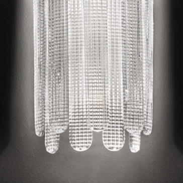 Contemporary Venetian Glass Wall Light A close-up view of the Contemporary Venetian Glass Wall Light reveals its textured, illuminated design. The fixture casts subtle shadows on a gray wall, highlighting the intricate grid pattern and curved edges of the glass.
