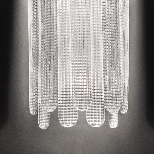 A close-up view of the Contemporary Venetian Glass Wall Light reveals its textured, illuminated design. The fixture casts subtle shadows on a gray wall, highlighting the intricate grid pattern and curved edges of the glass.