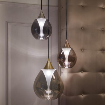 Three modern, teardrop-shaped pendant lights with golden tips hang in varying lengths against a beige, tufted wall. The lights have a glossy, translucent finish and emit a soft, warm glow, creating a cozy and elegant ambiance in the room.