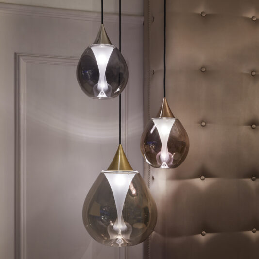 Exclusive Glass Pendant Light Set Three modern, teardrop-shaped pendant lights with golden tips hang in varying lengths against a beige, tufted wall. The lights have a glossy, translucent finish and emit a soft, warm glow, creating a cozy and elegant ambiance in the room.