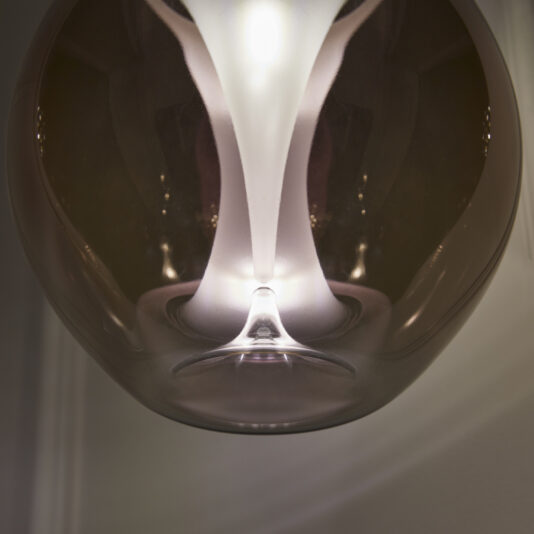 Exclusive Glass Pendant Light Cluster A close-up of a modern, spherical pendant light with a glossy, reflective surface. The light emits a warm glow, creating intricate reflections and patterns on the bulb’s surface. The background is blurred, focusing attention on the details of the light fixture.