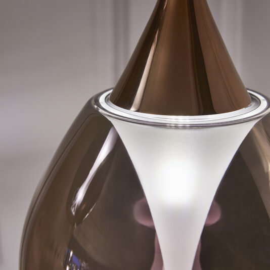 Exclusive Glass Pendant Light Cluster Close-up of a modern pendant lamp with a brown glossy exterior and a frosted white inner diffuser. The light is turned on, emitting a soft glow that illuminates the contours of the lamp. The background appears blurred, drawing focus to the lamp's design details.