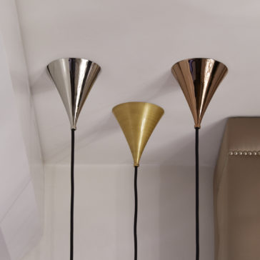 Three cone-shaped wall lamps are mounted on a white wall, positioned close to the ceiling. From left to right, they have metallic finishes in silver, gold, and copper. Each lamp has a black cord extending downward. The edge of a studded headboard is partially visible.