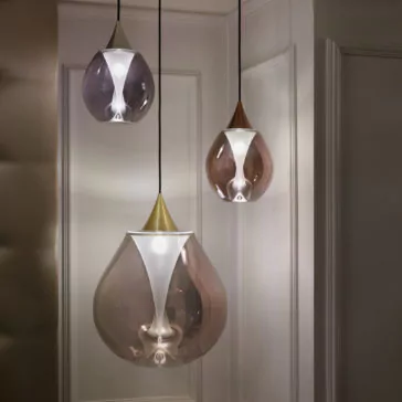 The Exclusive Glass Pendant Light Set, with its trio of teardrop-shaped, translucent shades in muted hues of violet, gold, and grey, hangs elegantly from the ceiling in a corner featuring white-paneled walls, casting a soft ambient glow.