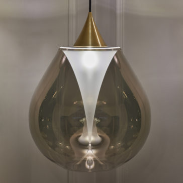 The Exclusive Glass Pendant Light Set showcases a teardrop-shaped, transparent glass shade. Within, the light is elegantly diffused by a frosted white cone and adorned with a brass or gold-colored cap. The design exudes sleek modern sophistication, hanging gracefully from a black cord.