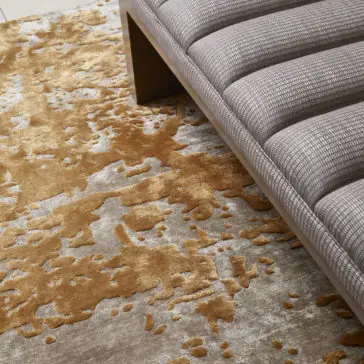 High Quality Abstract Hand Woven Rug A close-up view of a modern gray upholstered bench with a ribbed texture, placed on the High Quality Abstract Hand Woven Rug. The rug features a gold and beige design that resembles spilled or splattered paint. The setting appears minimalistic and sophisticated.