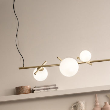 The Modern Murano Glass Horizontal Chandelier, featuring three spherical white bulbs, is elegantly suspended from the ceiling by a black wire and mounted on a sleek brass bar. Below it, a shelf displays books, decorative vases, and a small bowl against the backdrop of a plain beige wall.