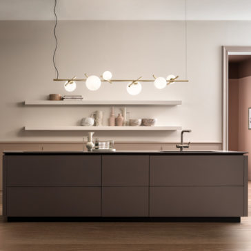 A modern kitchen with a large dark brown island counter is complemented by a Modern Murano Glass Horizontal Chandelier featuring round, white light fixtures suspended from the ceiling. The beige walls showcase two white floating shelves adorned with decorative items, while the island is equipped with a sink and faucet.