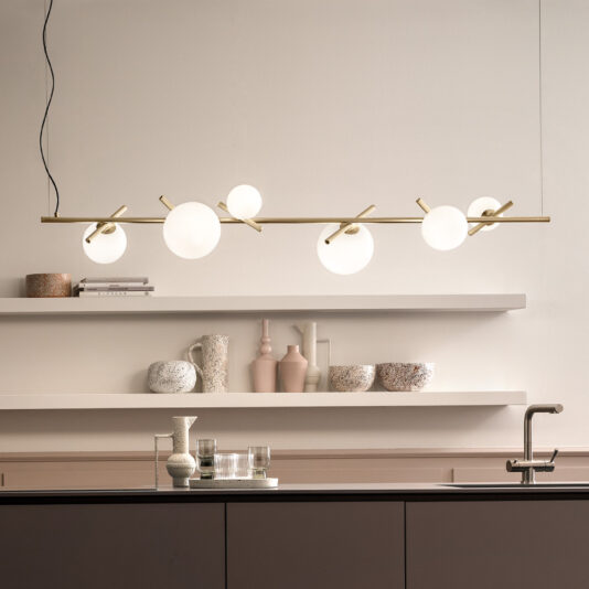 A modern kitchen with minimalist decor featuring the "Modern Murano Glass Horizontal Chandelier," showcasing six spherical white lights on elegant gold fixtures. The background includes floating white shelves adorned with various vases and decorative objects, alongside a countertop graced by a sleek faucet.