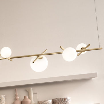 A Modern Murano Glass Horizontal Chandelier, featuring a sleek gold rod and three spherical white bulbs, hangs above a minimalist shelf adorned with various decorative items like a pink vase and textured objects. The backdrop is a light-colored wall.