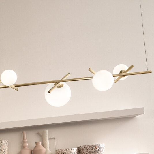 A Modern Murano Glass Horizontal Chandelier, featuring a sleek gold rod and three spherical white bulbs, hangs above a minimalist shelf adorned with various decorative items like a pink vase and textured objects. The backdrop is a light-colored wall.