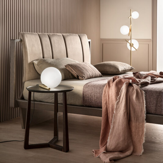 Modern Murano Glass Floor Lamp A contemporary bedroom showcases a wooden bed with beige upholstery, accented by beige and pink bedding. Beside the bed, a small round side table holds a spherical white lamp. In the corner, the Modern Murano Glass Floor Lamp, featuring three spherical lights, enhances the room's warm, neutral tones.