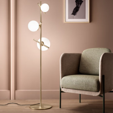 Modern Murano Glass Floor Lamp A modern interior showcasing a minimalist design. It features the Modern Murano Glass Floor Lamp, which has three spherical Murano glass lights on a sleek gold stand. Beside it is a stylish armchair upholstered in textured fabric with a green cushion. A framed abstract artwork adorns the wall behind them.