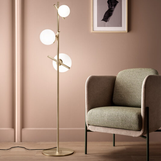 A modern interior showcasing a minimalist design. It features the Modern Murano Glass Floor Lamp, which has three spherical Murano glass lights on a sleek gold stand. Beside it is a stylish armchair upholstered in textured fabric with a green cushion. A framed abstract artwork adorns the wall behind them.