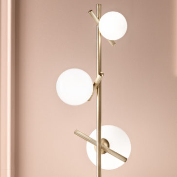 Modern Murano Glass Floor Lamp The Modern Murano Glass Floor Lamp boasts a gold metallic stand with three white spherical bulbs arranged at varying heights. Its sleek and contemporary design is elegantly showcased against a light pink wall.