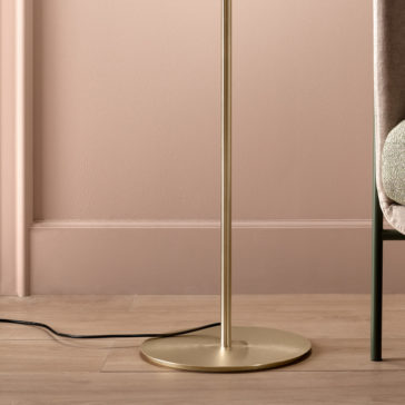 Modern Murano Glass Floor Lamp A close-up of the base of a Modern Murano Glass Floor Lamp, featuring a sleek, metallic gold stand. The lamp is placed next to a padded chair adorned with a textured cushion, while a black power cable lies on the light-colored wooden floor. The background wall is painted in soft pink tones.