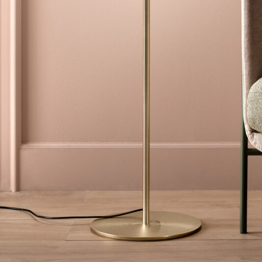 Modern Murano Glass Floor Lamp A close-up of the base of a Modern Murano Glass Floor Lamp, featuring a sleek, metallic gold stand. The lamp is placed next to a padded chair adorned with a textured cushion, while a black power cable lies on the light-colored wooden floor. The background wall is painted in soft pink tones.