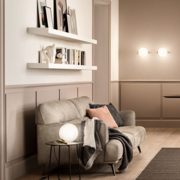 Modern Murano Glass Wall Light A cozy living room showcases a beige sofa adorned with pink and gray cushions, along with a draped gray blanket. Nestled beside the sofa is a small round side table featuring a white spherical lamp. Above, two white wall-mounted shelves exhibit books and framed pictures, softly illuminated by two circular Modern Murano Glass Wall Lights from the Modern Murano Glass Wall Light collection.