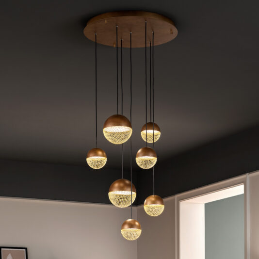 A Modern Pendant Drop Cluster Chandelier features six gold spherical lights suspended at varying lengths from a circular ceiling base, each with illuminated lower halves showcasing intricate patterns. The room highlights dark walls contrasted by a visible white doorway.