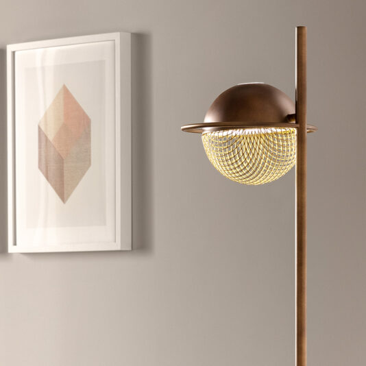 A Modern Round Floor Lamp with a bronze finish stands beside a framed geometric art print on a gray wall. The lamp features a globe-shaped, mesh-patterned light fixture with a partial dome top, casting a warm glow. The setting is minimalistic and contemporary.