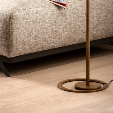 Modern Round Floor Lamp A Modern Round Floor Lamp with a circular base stands on a polished light wood floor beside a beige textured sofa. The sofa appears to have a black frame, and an open red book or magazine is visible on the seat.