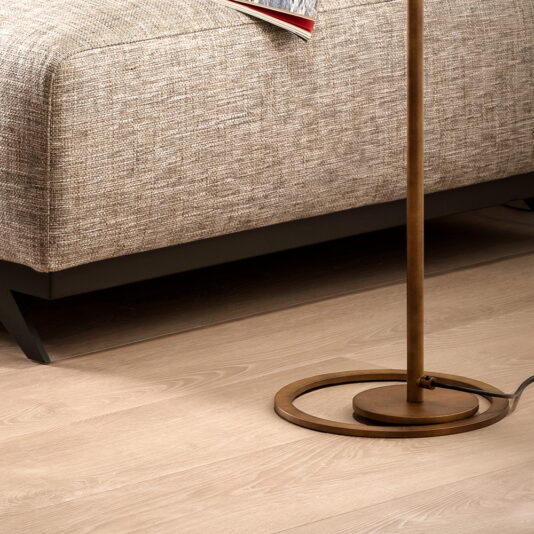 A Modern Round Floor Lamp with a circular base stands on a polished light wood floor beside a beige textured sofa. The sofa appears to have a black frame, and an open red book or magazine is visible on the seat.