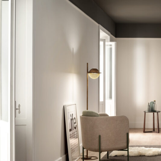 A minimalist living space featuring a beige armchair with green accents, positioned next to the Modern Round Floor Lamp. A framed print rests against the white wall. Sunlight filters in from a large window, illuminating a small wooden table with decorative items.