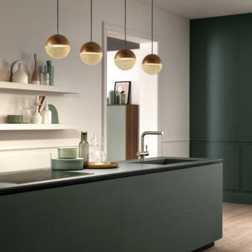 Modern kitchen with green cabinetry, black countertops, and gold spherical pendant lights. Minimalistic decor includes shelves with pastel-colored jars and vases. The sink island features a gold faucet and glassware. Walls in white and dark green shades complement the Modern Round Pendant Light design.