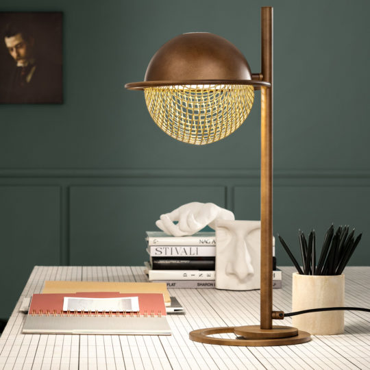 A Modern Round Table Lamp, featuring a stylish brown metallic frame and a spherical mesh shade that emits warm light, is placed on a gridded table. The table also holds a stack of books, a white face sculpture, and a container filled with black pencils. The background wall is painted green.