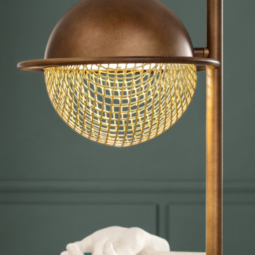 Modern Round Table Lamp Close-up of the Modern Round Table Lamp, featuring a modern, bronze-colored design with a dome-shaped shade and a textured, grid-patterned diffuser. The background showcases a green wall with subtle paneling, and the bottom of the image reveals part of a white object.