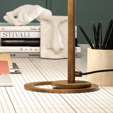 Modern Round Table Lamp A close-up of a stylish, modern desk featuring a sleek *Modern Round Table Lamp* with a bronze finish, a cluster of graphite pencils in a beige holder, a white sculpture of an eye and nose, and several stacked books. The desk has a white grid-patterned surface.