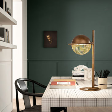 Modern Round Table Lamp A modern study desk with a tiled surface, adorned with the Modern Round Table Lamp, books, a notepad, and a pencil holder. A black chair is pulled up to the desk. The wall behind is a dark green with a framed picture, and white shelves on the left hold more books.