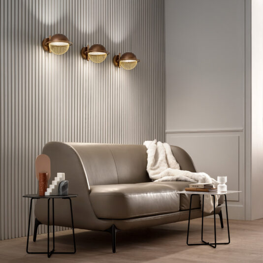Modern Round Wall Light A modern living room with a minimalist design. It boasts a sleek gray sofa adorned with a white blanket, accompanied by two small black metal side tables. The ambiance is enhanced by three brown and gold Modern Round Wall Lights. Vertical grooves on the wall add texture to the space.
