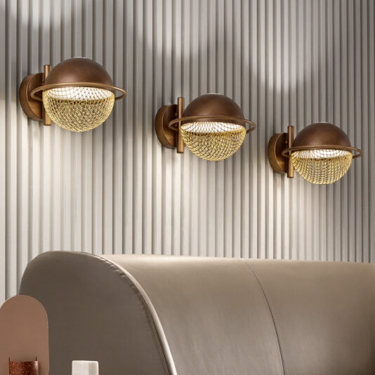 A modern interior features a curved, taupe couch in the foreground with three industrial-style wall sconces above. The sconces, made of bronze with netted glass covers, cast a warm glow against the vertical, fluted panel wall. The sleek **Modern Round Wall Light** nearby adds an elegant accent.