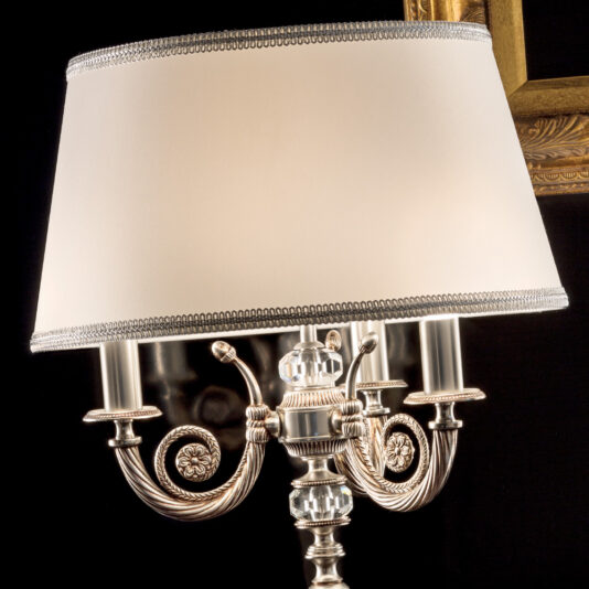 Classic Chrome Table Lamp A close-up of the Classic Chrome Table Lamp showcasing its white lampshade and intricately detailed metallic base adorned with ornate designs and glass accents. In the background, part of a gold picture frame is visible against a dark setting.