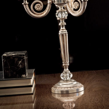 Classic Chrome Table Lamp A Classic Chrome Table Lamp stands beside an ornate silver candlestick holder with intricate designs on a polished wooden surface. Three books, topped with a marbled block, complete the arrangement against a dark background. The entire scene reflects sophistication and elegance.