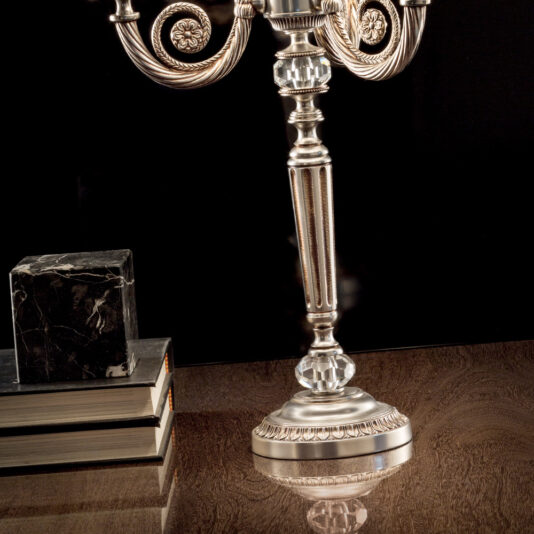 Classic Chrome Table Lamp A Classic Chrome Table Lamp stands beside an ornate silver candlestick holder with intricate designs on a polished wooden surface. Three books, topped with a marbled block, complete the arrangement against a dark background. The entire scene reflects sophistication and elegance.