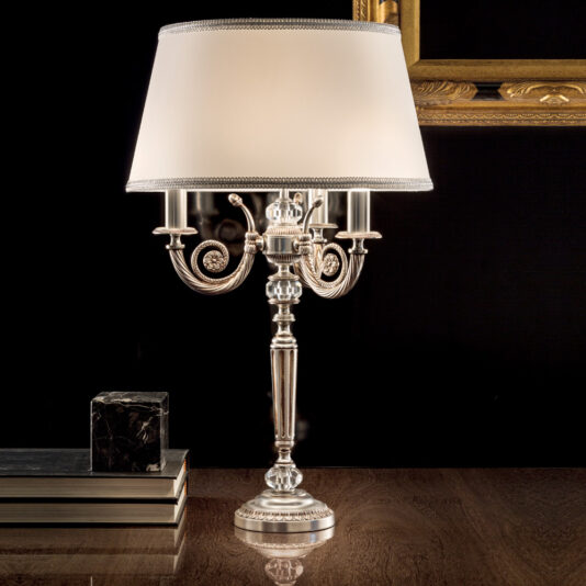 The Classic Chrome Table Lamp, with its ornate base featuring three curved arms and a white fabric lampshade, sits on a wooden surface next to a stack of books and a small decorative black and silver box. A gilded picture frame is partially visible in the background.