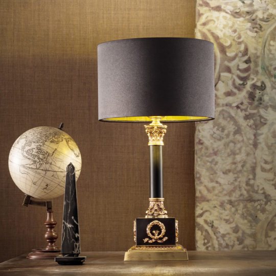 A Classic Louis Table Lamp adorned with a black shade and intricate gold detailing stands on a wooden surface. Beside it, you'll find an antique-style globe resting on a wooden stand and an obelisk-shaped ornament made of black marble. The backdrop features textured, patterned wallpaper.