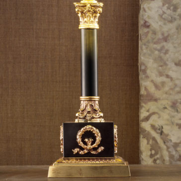 Classic Louis Table Lamp An ornate, tall ornamental pillar, reminiscent of a Classic Louis Table Lamp, made of dark stone and gold with intricate gold leaf detailing at the base and top. It stands on a rectangular dark base accented with gold laurel wreaths and is set against a brown marbled background.