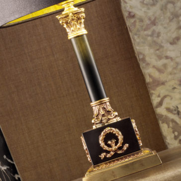Classic Louis Table Lamp A close-up of the Classic Louis Table Lamp reveals its detailed gold base adorned with intricate leaf patterns and accented by a decorative wreath design on a black stand. The background showcases a marble texture alongside a rich brown fabric surface.