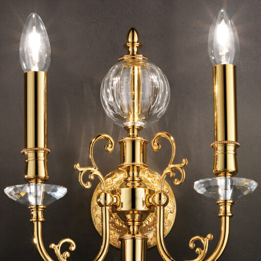The Classic Transparent Gold Wall Light is a striking, golden wall sconce adorned with intricate detailing, featuring two candle-like light bulbs positioned on either side of a transparent glass orb. This ornate fixture showcases elaborate curving elements and diamond-shaped crystal accents beneath each bulb, all mounted against a dark background.