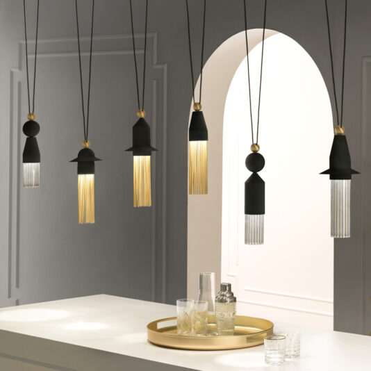 A modern interior showcases a Contemporary Black LED Pendant Light Cluster arrangement, elegantly suspended in a row above the white countertop. Each pendant light boasts a distinct design, combining black, gold, and glass elements. On the countertop sits an exquisite gold tray with glassware, while a white arched doorway serves as the backdrop.