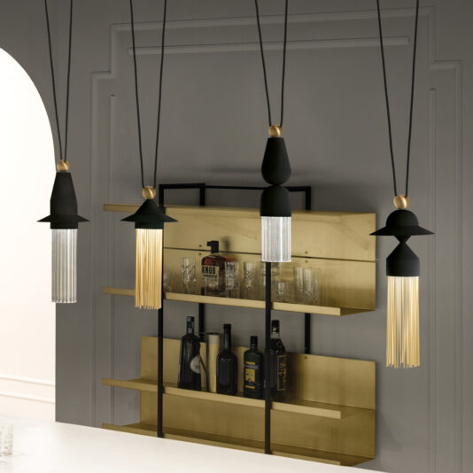 A modern interior space featuring a **Contemporary Black LED Pendant Light Cluster** with gold accents hanging over a white counter. The background showcases a gold and black shelving unit with various bottles and glasses neatly arranged. The walls are painted in a light gray color.