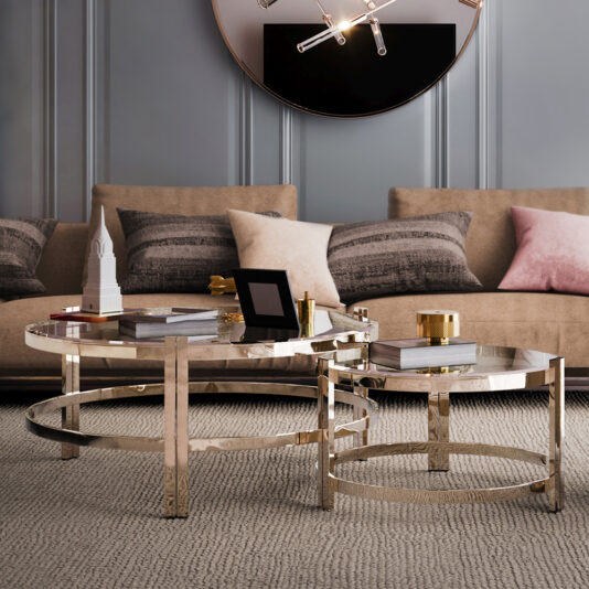 A contemporary luxury living room features a beige sofa accented with grey and pink pillows. In front of the sofa are two Contemporary Luxury Glass Coffee Tables with gold-colored frames, adorned with decorative items including a small model building, books, and trays.