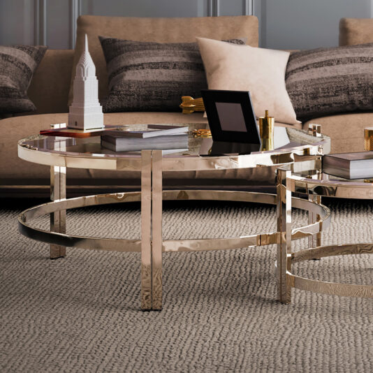 A sophisticated living room features a beige sofa adorned with patterned pillows. In front stands a sleek, contemporary coffee table from the Contemporary Luxury Glass Coffee Tables collection, featuring elegant gold frame details. The table displays decorative items such as a picture frame, books, and a miniature building model on top of a textured rug.