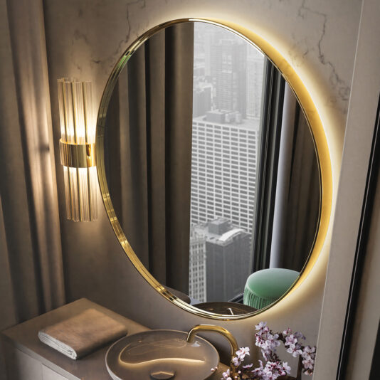 The Contemporary Round Mirror, featuring a gold frame and backlighting, highlights a stylish bathroom vanity. An oval sink with an elegant gold faucet sits next to a neatly folded towel. A vase filled with pink flowers adds charm to the countertop, while the mirror captures a stunning reflection of a cityscape with tall buildings.