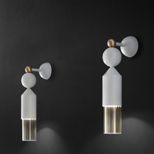 A Contemporary White LED Wall Light is mounted on a dark wall. The light features geometric shapes, including a round brass element, white circular and cylindrical sections, and a bottom part with vertically aligned transparent tubes that cast a soft downward glow.