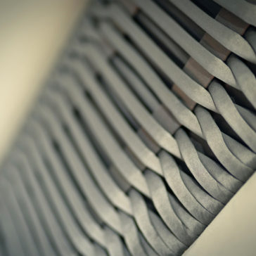 A close-up image of the Designer Velvet And Satin Interlaced Bed showcases tightly woven gray fabric strips arranged in an intricate lattice pattern. The strips, which are reminiscent of designer velvet and satin intricately interlaced with care, overlap each other at uniform intervals, creating a three-dimensional braided appearance. The lighting highlights the depth and detail of the weave.