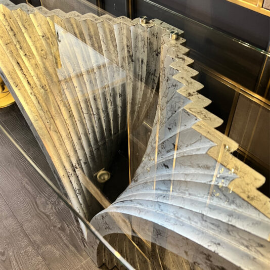 Exclusive Contemporary Glass Console Table A glass table with a distinct, spiraling gray marble base featuring a zigzag pattern rests on a dark wooden floor. The Exclusive Contemporary Glass Console Table showcases an intricate design that adds an eye-catching, modern aesthetic.
